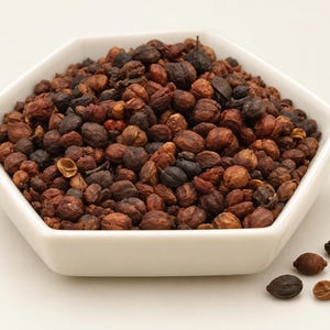 May include: A white hexagonal bowl filled with a mound of dried, dark brown, textured seeds. The seeds vary in shade from light tan to deep blackish-brown, featuring a wrinkled, irregular surface. A few loose seeds are scattered near the bowl.