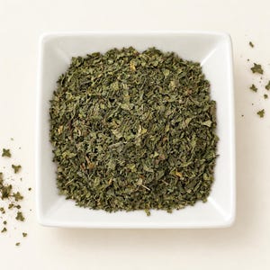 May include: A square white ceramic bowl filled with a mound of dried, finely chopped dark green herbs. The texture of the herbs is coarse and flaky, with varying shades of olive and forest green visible throughout the pile of dried leaves.