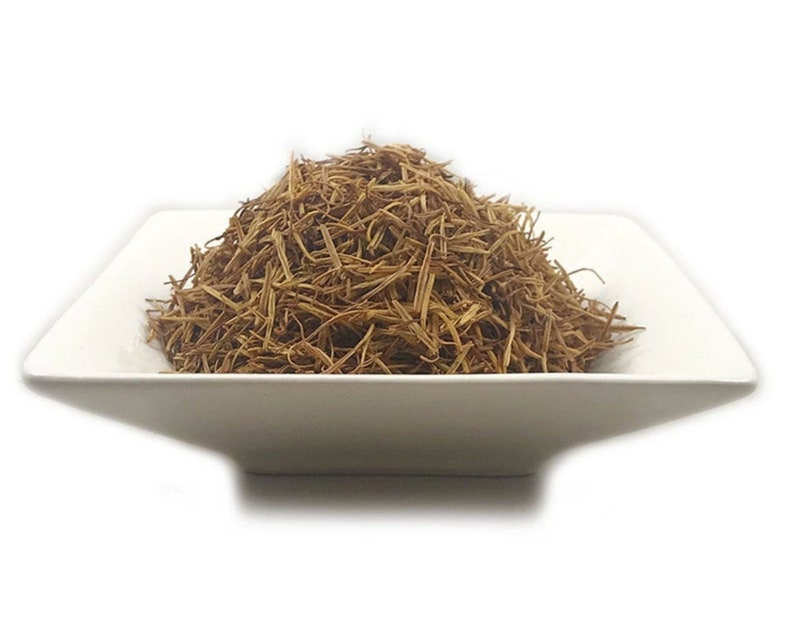 May include: A mound of dried, thin, light brown herbal stems and fibrous plant material piled high in a square, white ceramic bowl. The texture of the botanical pieces is shredded and brittle, creating a dense, textured heap of organic matter.