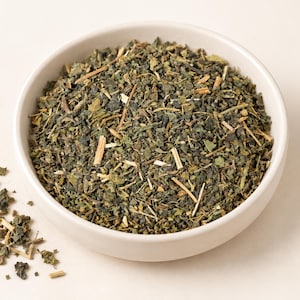 May include: A white ceramic bowl filled with a mixture of dried green leaves and small light brown woody stems. The herbal blend consists of finely chopped dark green foliage and thin, rigid plant stalks, with a small pile of the same mixture scattered outside the bowl.