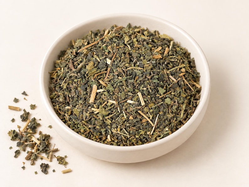May include: A white ceramic bowl filled with a mixture of dried green leaves and small light brown woody stems. The herbal blend consists of finely chopped dark green foliage and thin, rigid plant stalks, with a small pile of the same mixture scattered outside the bowl.