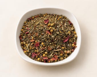 May include: A white bowl filled with a loose-leaf herbal tea blend. The mixture contains dried green herbs, small yellow chamomile flowers, and vibrant pink rose petals, creating a textured, multi-coloured botanical composition.