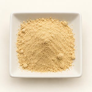 May include: A square white ceramic bowl filled with a mound of fine, light beige powder. The powder has a dry, granular texture with small clumps scattered throughout the pile, centred within the bowl against a plain, neutral background.