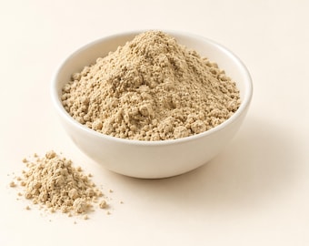 May include: A white ceramic bowl filled with a mound of fine, light beige powder. The powder has a dry, granular texture and is piled high in the center of the bowl, with a small additional pile of the same beige substance resting nearby.