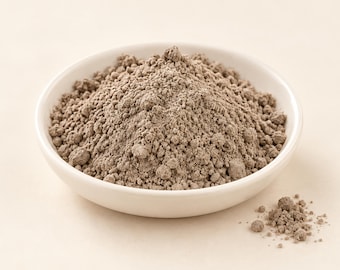 May include: A small, round, white ceramic bowl filled with a mound of fine, dusty, light brown powder. The texture of the powder appears granular and dry, with a few small clumps scattered throughout the pile and a small amount spilled outside.
