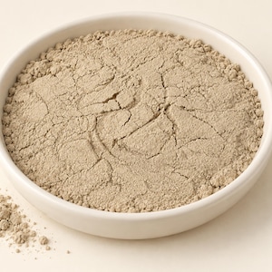 May include: A round white ceramic bowl filled with a fine, light beige powder. The surface of the powder shows a network of dry, cracked fissures across its texture. A small amount of the same beige powder is scattered outside the bowl.