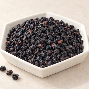 Könnte beinhalten: Eine weiße, sechseckige Schale, gefüllt mit einem Haufen getrockneter, dunkelvioletter bis schwarzer Beeren. Die Oberfläche der Beeren wirkt faltig und strukturiert. Drei einzelne getrocknete Beeren liegen separat außerhalb der Schale unten links.