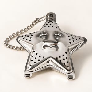 May include: A star-shaped stainless steel tea infuser featuring a molded face with eyes, a nose, and a mouth. The metal surface is perforated with small circular holes for steeping tea leaves and includes an attached silver-toned metal chain.