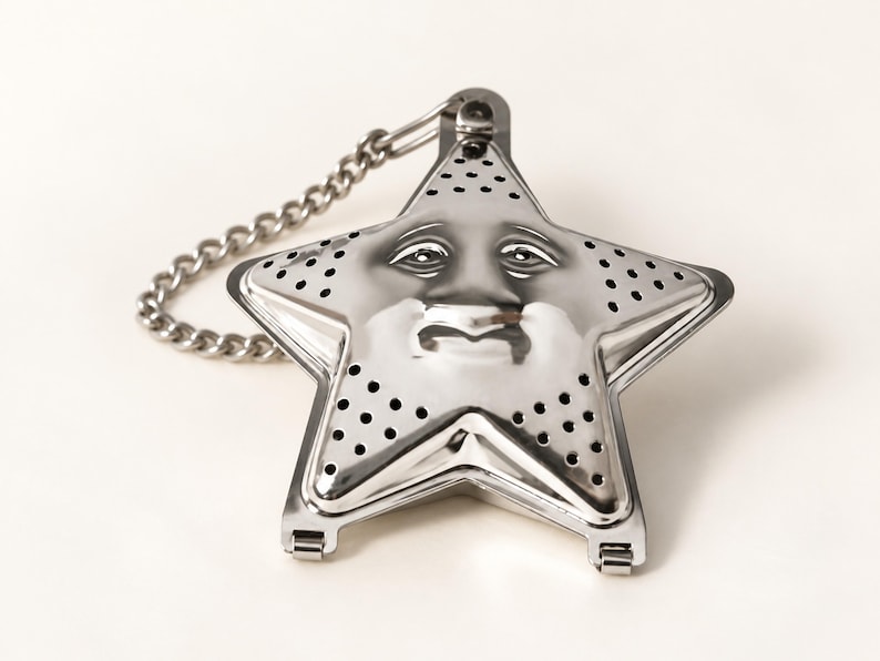 May include: A star-shaped stainless steel tea infuser featuring a molded face with eyes, a nose, and a mouth. The metal surface is perforated with small circular holes for steeping tea leaves and includes an attached silver-toned metal chain.