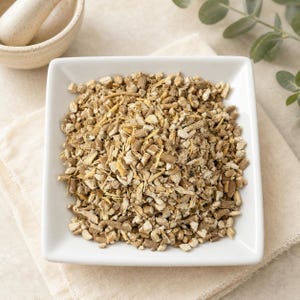 May include: A square white ceramic bowl filled with a pile of dried, chopped light brown and beige botanical root pieces. The texture of the dried plant material is coarse and irregular, featuring a mix of woody fragments and fibrous strands.