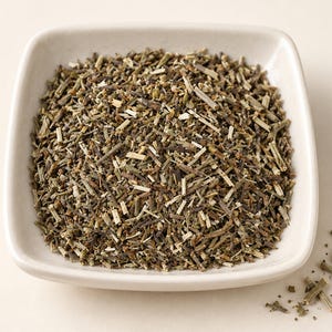 May include: A square, off-white ceramic bowl filled with a mound of dried, chopped green and brown herbal plant matter. The mixture consists of small, thin, woody stems and fragmented leaves, with a few loose pieces scattered outside the bowl.