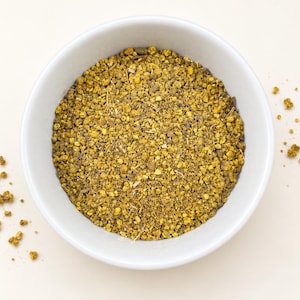 Könnte beinhalten: Eine weiße Keramikschale gefüllt mit einer Mischung aus kleinen, körnigen gelben und braunen Bienenpollenpellets. Die Textur ist grob und ungleichmäßig und besteht aus winzigen, unregelmäßigen Partikeln und dünnen Pflanzenfragmenten.