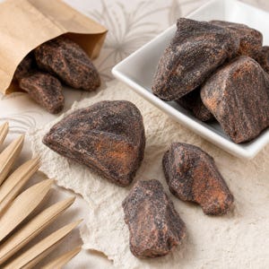 May include: Several dark brown, irregularly shaped, textured chunks of raw cacao butter or chocolate pieces. Some pieces are contained within a white square bowl and a brown paper bag, while others are scattered on a textured off-white surface.