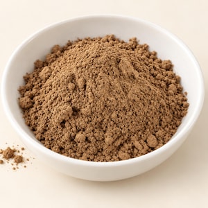 May include: A white ceramic bowl filled with a mound of fine, light brown powder. The texture of the powder appears dry and granular, with a few small loose particles scattered outside the bowl near the bottom left edge of the container.