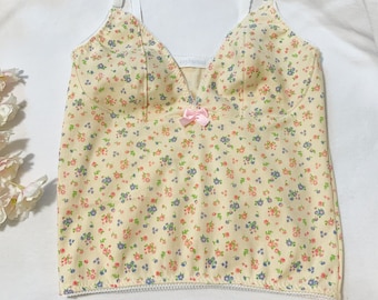 May include: A yellow floral print tank top with thin white straps and a small pink bow at the center. The top has a scalloped hem.