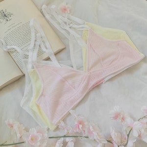 May include: Three layered bras in white, yellow, and pink with delicate lace trim. The bras are arranged on a white surface with a book and pink flowers.