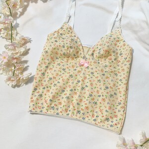 May include: A yellow floral print spaghetti strap top with a small pink bow at the center. The top has a white trim around the neckline and bottom hem.