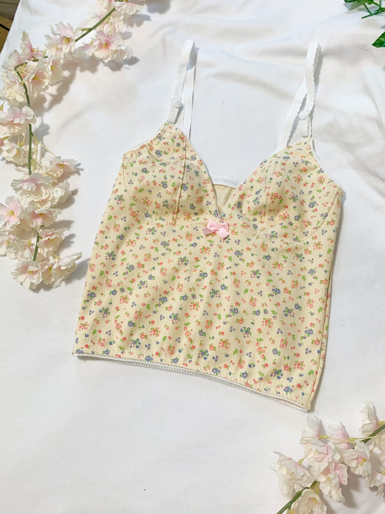 May include: A yellow floral print spaghetti strap top with a small pink bow at the center. The top has a white trim around the neckline and bottom hem.