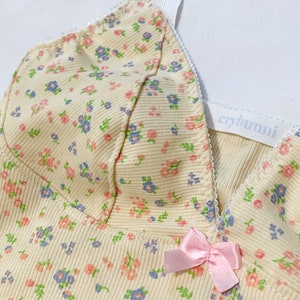 May include: A yellow ribbed tank top with a floral print and a pink bow. The tank top has adjustable straps and a white label that says "crybuni".