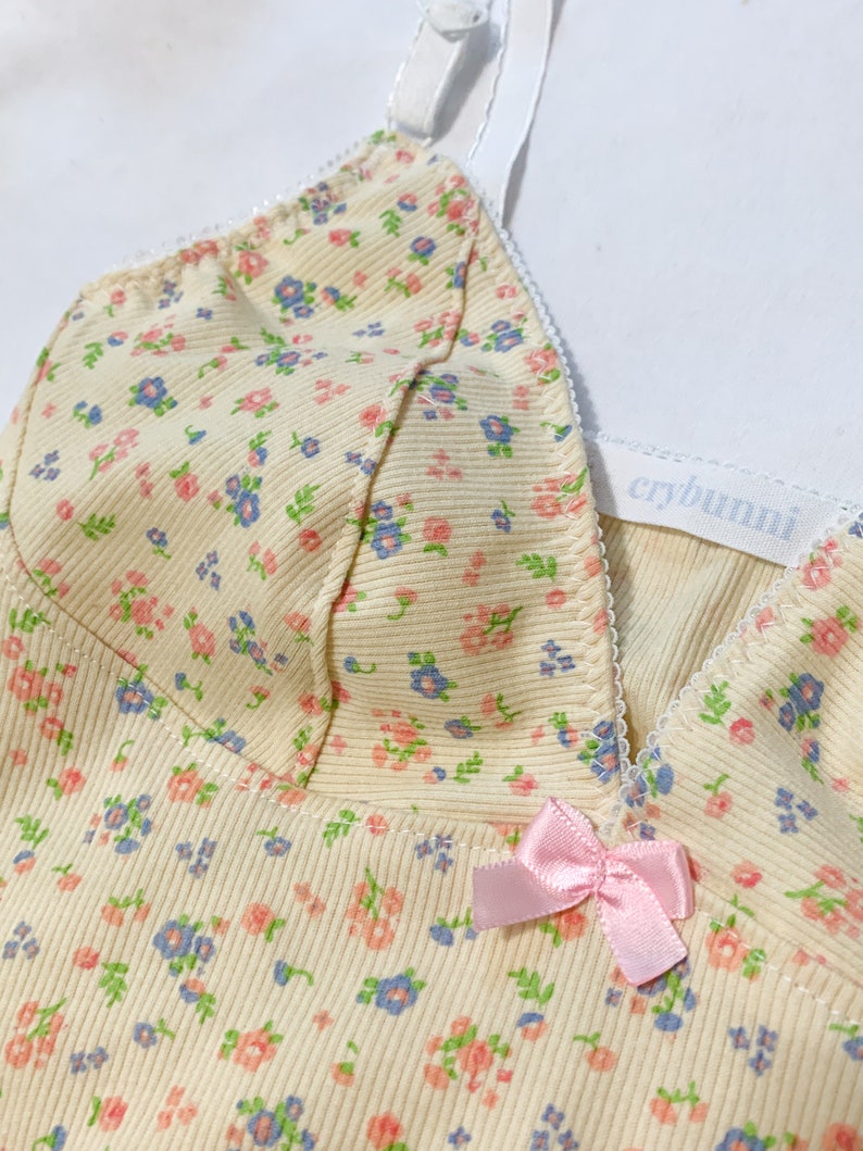 May include: A yellow ribbed tank top with a floral print and a pink bow. The tank top has adjustable straps and a white label that says "crybuni".