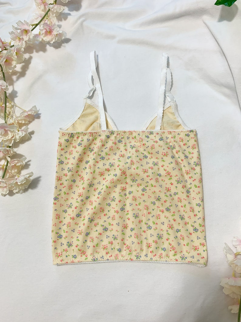 May include: A yellow tank top with a white floral print and white straps. The tank top has a scalloped hem.