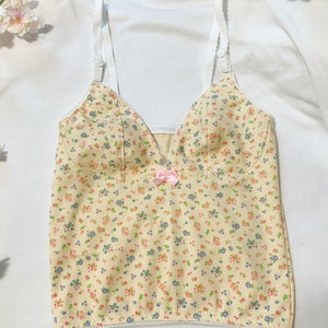 May include: A yellow tank top with a floral print and a pink bow. The tank top has white straps and a white trim around the bottom.