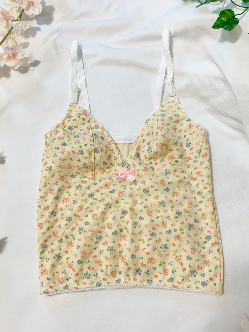 May include: A yellow tank top with a floral print and a pink bow. The tank top has white straps and a white trim around the bottom.