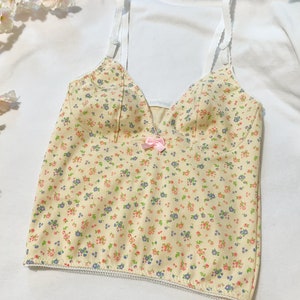 May include: A yellow floral print tank top with thin white straps and a pink bow at the center. The top has a scalloped hem and a ribbed texture.