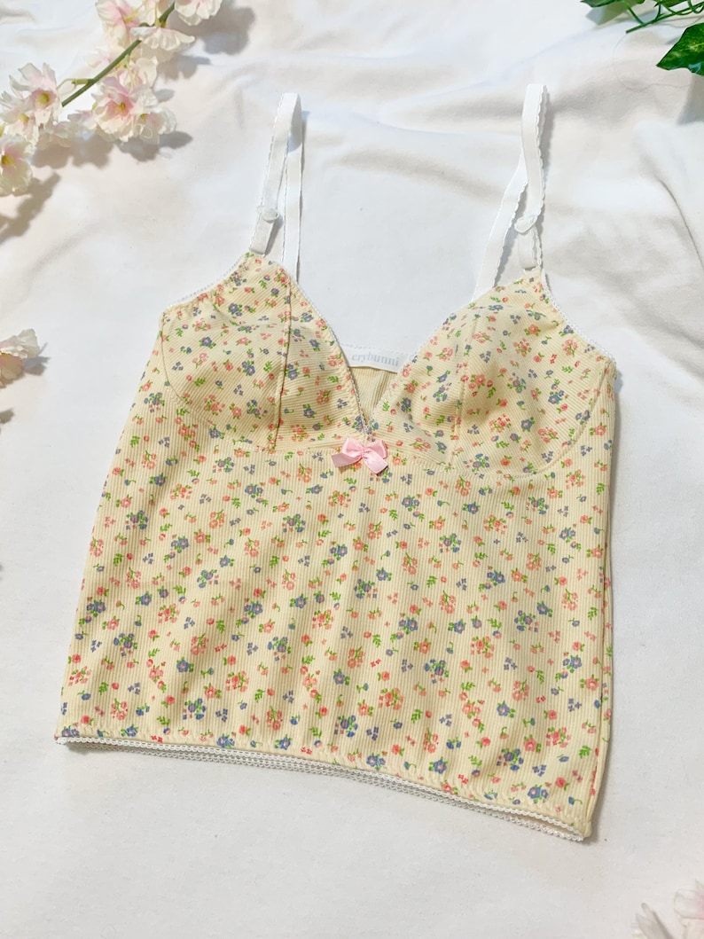 May include: A yellow floral print tank top with thin white straps and a pink bow at the center. The top has a scalloped hem and a ribbed texture.