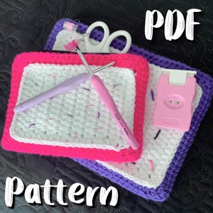 May include: A crochet pattern with the text "PDF" and "Pattern". The image shows a white and pink crochet square, a pink and purple crochet pouch, two crochet hooks, a pair of scissors, and a pink row counter.