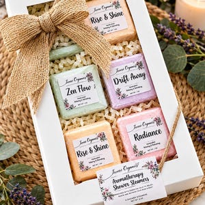 May include: A white gift box containing six square shower steamers in peach, light green, lavender, and pink. Each steamer has a label reading Rise & Shine, Zen Flow, Drift Away, or Radiance. The box is decorated with a large burlap bow and a tag.