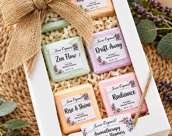 May include: A white gift box containing six square shower steamers in orange, green, purple, and pink. Each steamer has a label reading Ivoré Organics with names like Rise & Shine, Zen Flow, Drift Away, and Radiance. A burlap bow decorates the box.