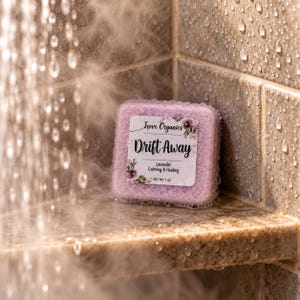 May include: A square, light purple shower steamer with a white label reading Trove Organics Drift Away Lavender Calming & Healing NET WT 28g. The product has a textured, porous surface and is covered in water droplets in a shower setting.