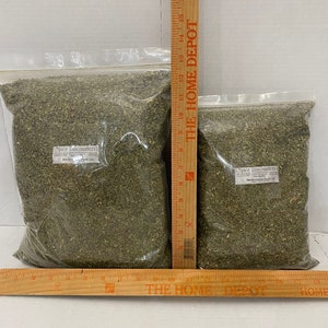 May include: Two clear plastic bags filled with dried green herbs labeled "Spice Discounters" with the text "Marjoram Herbs 1 oz" on the label. The bags are next to a wooden ruler with the text "THE HOME DEPOT" printed on it.