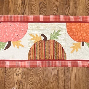 May include: A red, orange, and pink patchwork table runner with a fall theme. The runner features four pumpkins with green leaves and a red and white plaid border.