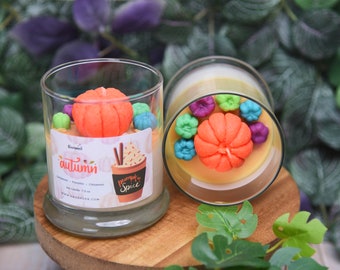 May include: Two clear glass jar candles with orange pumpkin-shaped wax toppers and smaller green, blue, and purple pumpkin accents. The front label features the text autumn and pumpkin spice with an illustration of a drink in a cup.