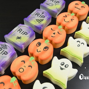May include: A collection of Halloween-themed bath bombs arranged in rows, featuring purple and green tombstone shapes with RIP text, orange pumpkins with various facial expressions, and white ghosts with green-tinted bases and Oavender text.