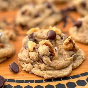 May include: A close-up of a wooden serving tray with a black honeycomb pattern around the edge. The tray is filled with chocolate chip cookies with pecans and sea salt.
