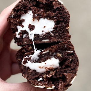 Puede incluir: Una sección transversal de un postre de chocolate revela un centro de malvavisco blanco. El exterior marrón oscuro está salpicado de nueces y chispas de chocolate. Parte del malvavisco gotea por el costado.