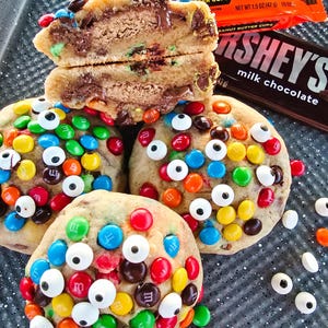 Op de afbeelding: Close-up van verschillende versierde koekjes. Eén koekje is doormidden gesneden en onthult een vulling van chocolade en pindakaas. De andere koekjes zijn rond en bedekt met kleurrijke snoepjes en eetbare snoepogen. Reese's Peanut Butter Cups en een Hershey's melkchocoladereep zijn op de achtergrond.