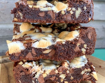 Puede incluir: Una pila de cuatro brownies de chocolate cubiertos con malvaviscos, chispas de chocolate y nueces. Los brownies están dispuestos sobre una tabla de cortar de madera.