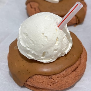 Puede incluir: Primer plano de un postre que se asemeja a un batido. Una base de galleta de color rojo está cubierta con un glaseado marrón y una gran bola de crema batida blanca. Una pajita a rayas rojas y blancas está insertada en la crema.