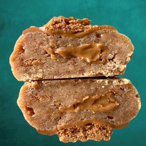 May include: A close-up of a split cookie with a brown filling. The cookie is made with a light brown dough and has a crispy texture. The filling is a smooth, creamy peanut butter.
