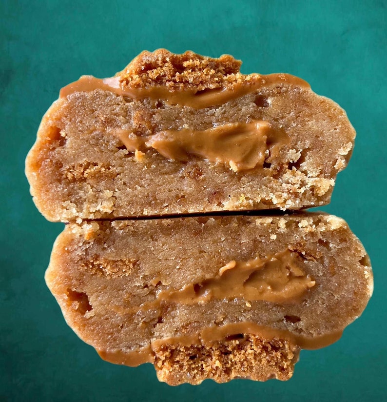 May include: A close-up of a split cookie with a brown filling. The cookie is made with a light brown dough and has a crispy texture. The filling is a smooth, creamy peanut butter.
