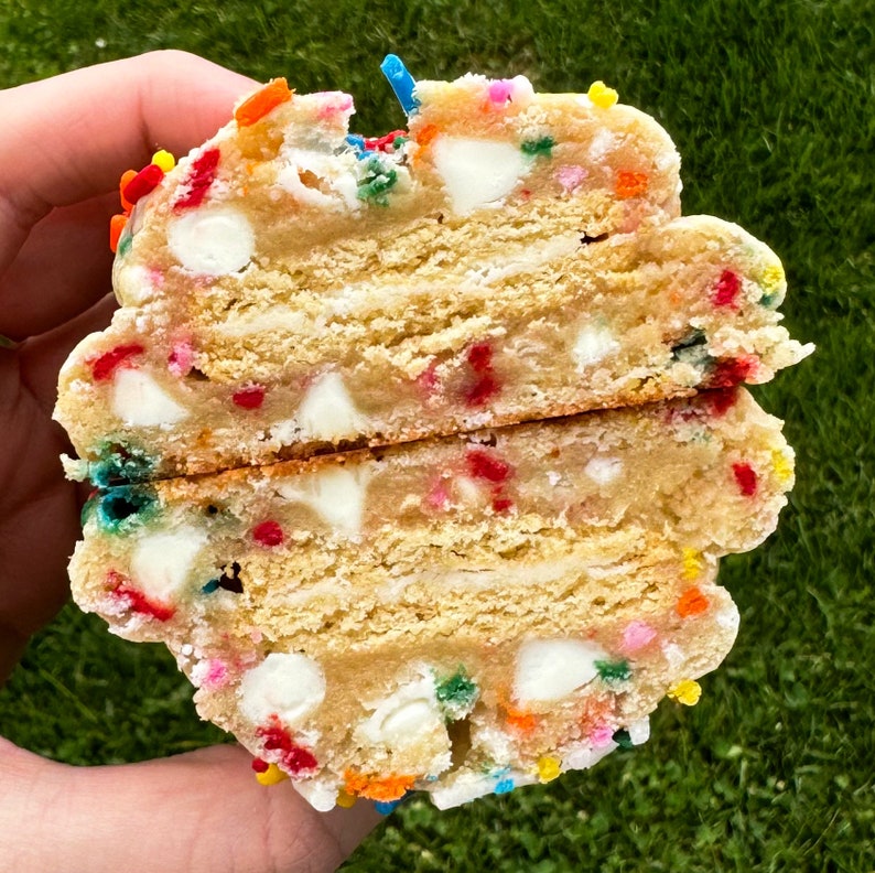 Puede incluir: Primer plano de una galleta grande y redonda con chispas de chocolate blanco y espolvoreado de colores. La galleta est&aacute; cortada por la mitad, revelando capas de masa y chocolate blanco.