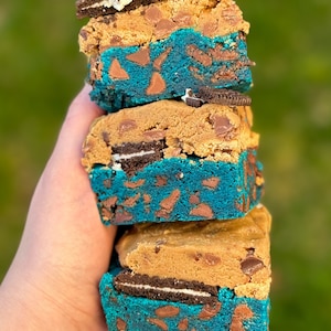 May include: A stack of four blue and brown cookie bars with Oreo cookies inside. The bars are held in a hand.