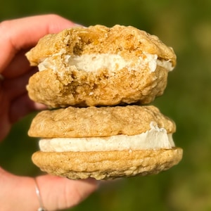 May include: Two oatmeal cookies with a creamy white filling. The cookies are stacked on top of each other, and the top cookie is slightly broken.