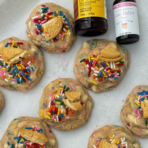 Puede incluir: Primer plano de seis galletas de masa para pastel con chispas de colores y trozos de galletas Oreo en la parte superior. Dos botellas de saborizante son visibles en el fondo, una etiquetada "Butter Vanilla Bakery Emulsion" y la otra etiquetada "Cake Batter Flavor".