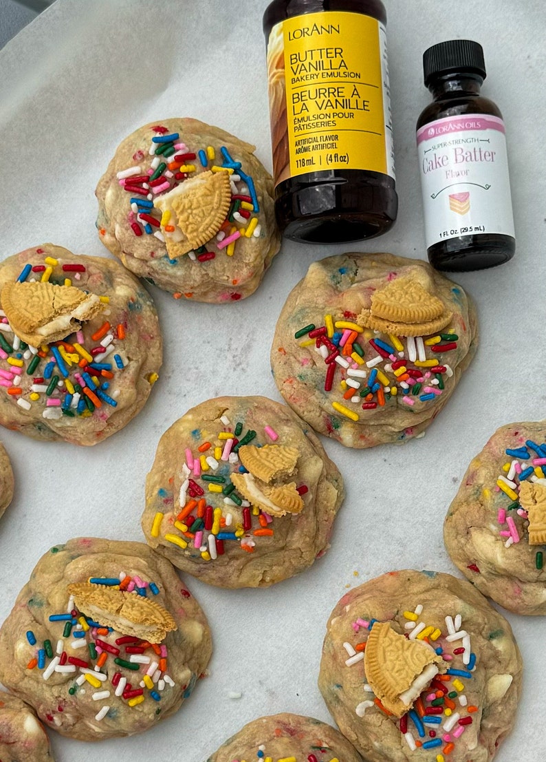 Puede incluir: Primer plano de seis galletas de masa para pastel con chispas de colores y trozos de galletas Oreo en la parte superior. Dos botellas de saborizante son visibles en el fondo, una etiquetada "Butter Vanilla Bakery Emulsion" y la otra etiquetada "Cake Batter Flavor".