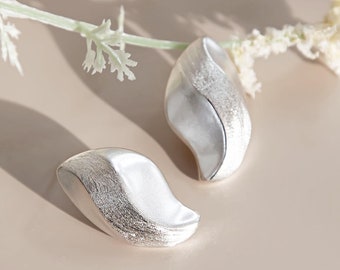 May include: A pair of silver earrings shaped like a leaf with a textured surface.